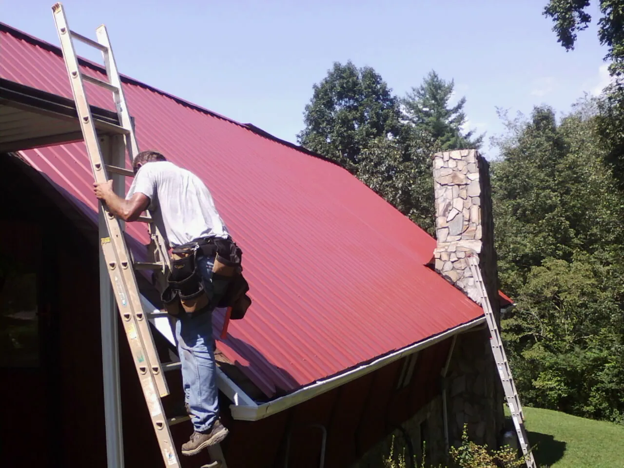 Roofer on a ladder beside a red metal roof and stone chimney — Soffit & Fascia Repair by Roofing Brothers in D Lo