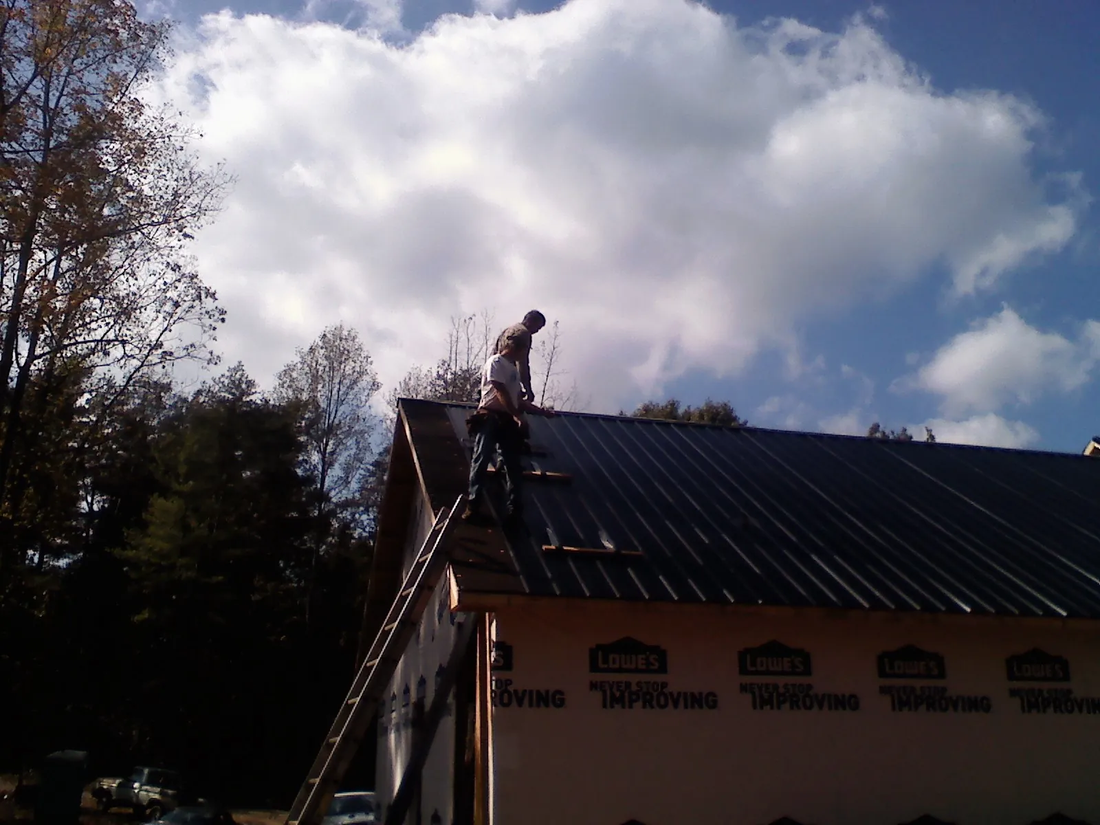 Roofer fastening dark metal panels high against a cloudy sky, from a Roof Inspection job in D Lo